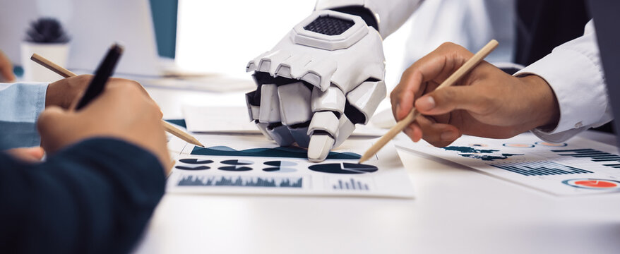 A human and a robotic hand engage in a collaborative business meeting, analyzing data charts together, illustrating technology's role in innovative teamwork.Noogenesis
