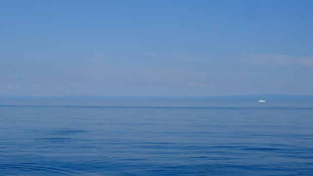 Alone sailboat floating in vast open sea under serene blue sky, tranquil ethereal view. Waves calmly washing up rocky sea beach. Stone skipping across a calm ocean. Tranquil and spiritual seascape
