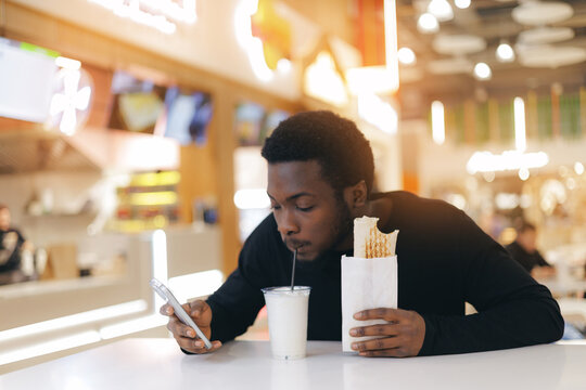 Young african male use phone and enjoying wrap and drink in food court while browsing phone