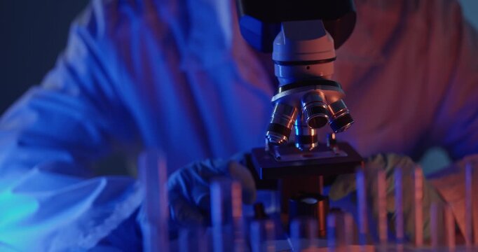 Microscope scientist laboratory focus adjustment closeup of gloved hands rotating objective and sliding sample while test tubes shift under blue neon lighting.