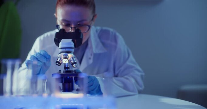 Female scientist in biotech lab adjusting microscope and focusing on sample with gloved hands.