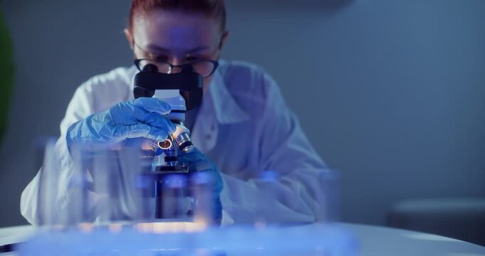 Female scientist microscope laboratory focusing on moving sample through eyepiece; gloved hands adjust controls in blue lit clinical research closeup.