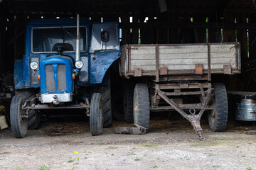 Obraz premium Old Blue Tractor And Wooden Trailer Parked In Rustic Barn On Rural Farm