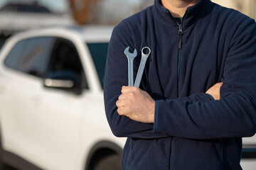 Roadside assistance. Male auto mechanic stands with crossed arms holding wrenches, parked vehicle visible in the background under natural lighting conditions © Kateryna Muzhevska
