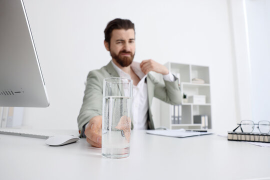 Man with glass of water suffering from heat at table near white wall indoors, selective focus