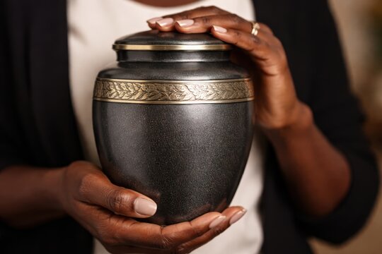 African American woman hands holding dark metal cremation urn with ashes.
