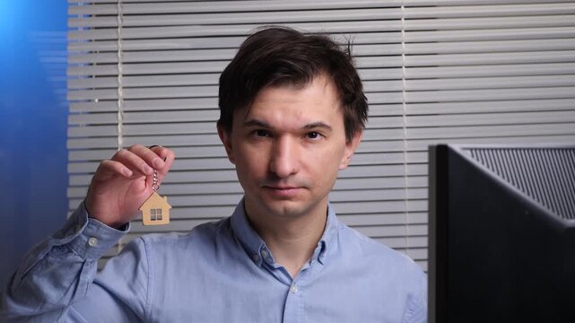 Man holding a tiny house shaped wooden keychain, symbolizing home ownership, real estate investment, and property dreams against a blurred office background