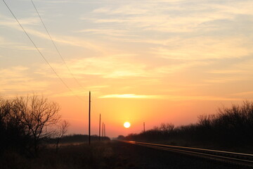 Sunset over rural railroad tracks with glowing horizon, power lines and silhouetted trees creating strong leading lines in warm countryside evening light.