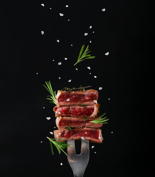 Beef pieces on meat fork, thyme, rosemary and salt against black background