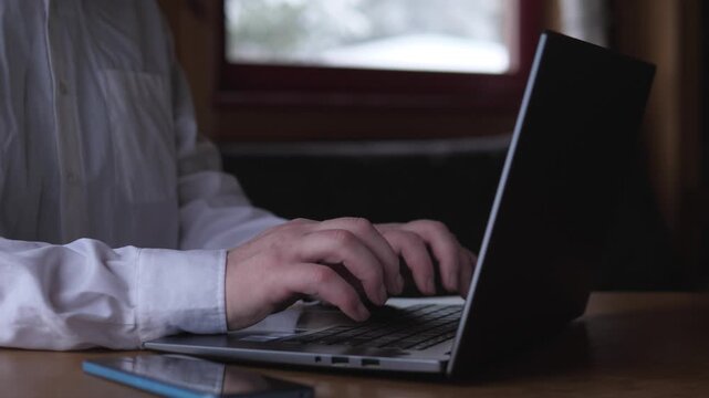 Closeup Hands Typing On Laptop In Dim Home Office By Window, Freelancer Composing Email And Documents, Smartphone And Notebook On Wooden Desk, Warm Moody Lighting And Calm Focused