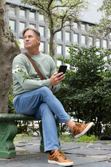 Obraz premium Middle-aged man sitting on green bench in urban plaza holding phone and white flowers in sweatshirt