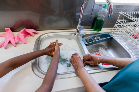 African senior and youth women washing dishes together at kitchen sink with soapy water and sponge