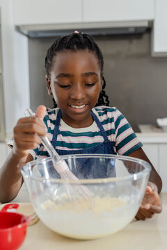 African American denim-aproned child mixing batter with whisk in glass bowl at counter by red cup