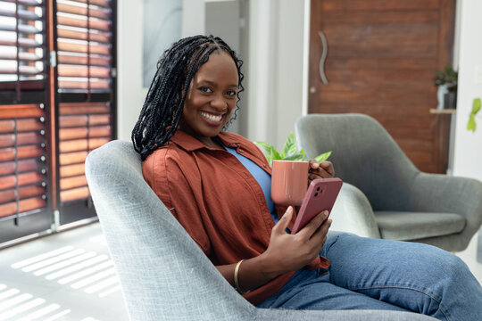 Adult woman sitting in armchair at home near shutters, holding pink mug, phone, wearing rust shirt