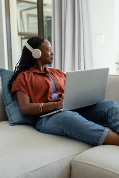 Adult African woman sitting on couch near window wearing rust top, jeans headphones holding laptop