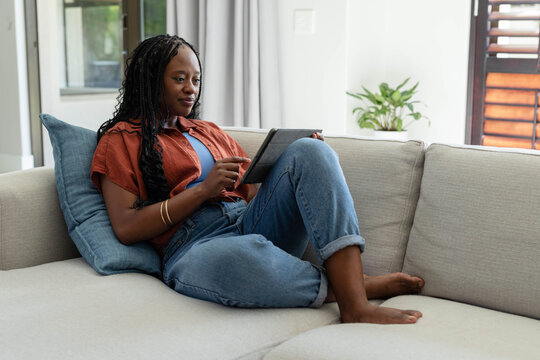 Adult African woman reclining on sofa holding tablet, wearing rust shirt and jeans at home