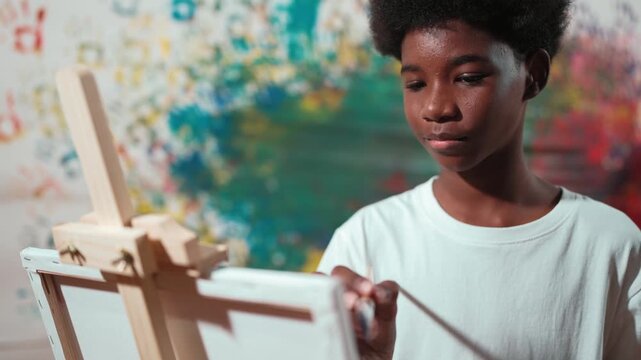 African boy paint blue water color by using paint brush to canvas in art lesson. high school student drawing or painting to express creativity while standing at colorful stained wall. Edification