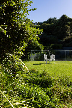Ornate white chairs are facing each other with round table on manicured lawn near glass fence