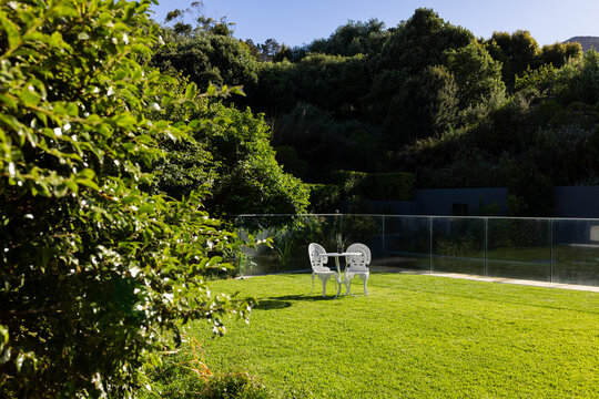 White bistro table and two chairs sitting in garden, glass railing, sunlight casting shadows