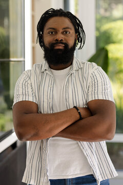 Man with dreadlocks standing arms crossed by glass doors, wearing striped shirt and beaded bracelet