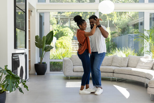 African American couple dancing, holding hands, wearing jeans, sneakers in living room with windows