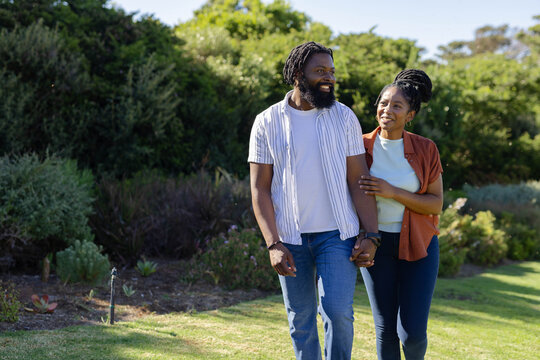Couple holding hands walking in park, man with dreadlocks, woman with braided bun near sprinkler