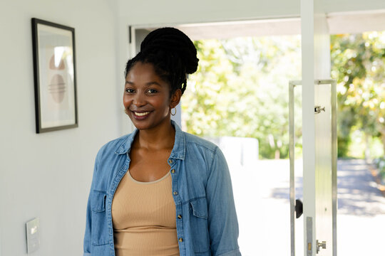 Woman standing in bright home entryway near open front door, wearing denim shirt and hoop earrings