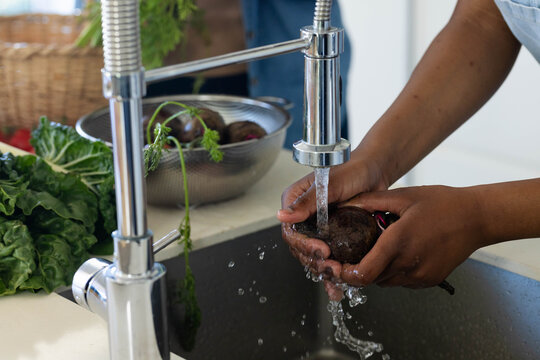 Hands rinsing dark beet under chrome pull-down faucet at kitchen sink, colander and greens