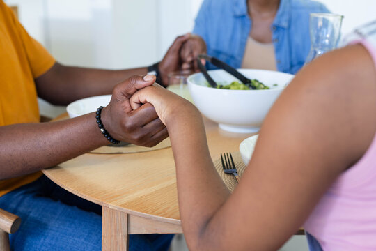 African American family holding hands and sharing salad at round dining table with black tongs
