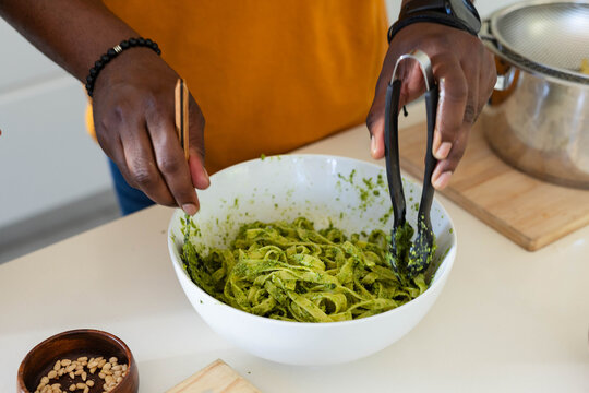 White bowl of pesto fettuccine being stirred with black silicone tongs and fork on countertop