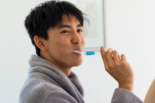 Asian man wearing gray bathrobe brushing teeth at home with clear toothbrush and toothpaste foam