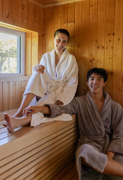 Couple sitting on wooden sauna benches wearing white and gray bathrobes, smiling toward camera