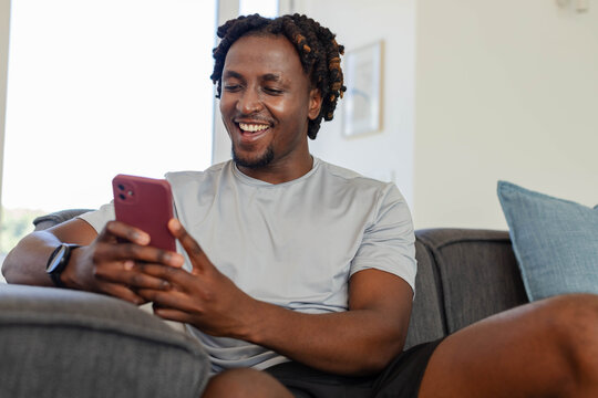 African American man sitting on gray sofa in living room holding pink phone and checking smartwatch