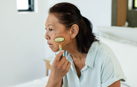 Senior Asian woman sitting on bed, wearing light blue collared shirt, rolling jade facial roller
