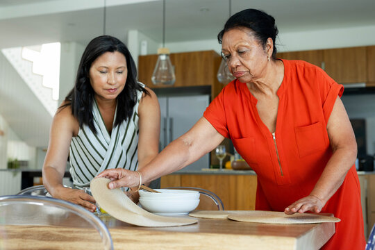 African American women, one senior in orange dress, arranging woven mats on wood table in kitchen