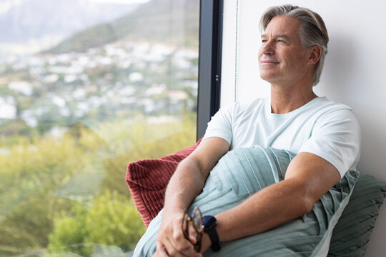 Middle-aged man sitting by floor-to-ceiling window holding glasses and pleated cushion, copy space