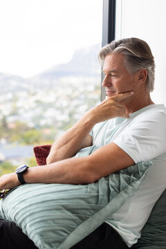 Senior man wearing tee sitting on couch by window holding cushion, glasses and watch, copy space