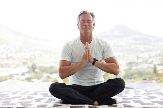 Mature man sitting cross-legged on patterned-mat by tall window praying wearing tee and smartwatch