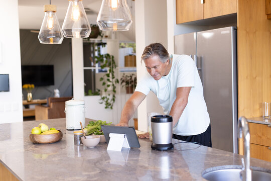 Senior man standing at kitchen island leaning on counter looking at tablet on stand, copy space