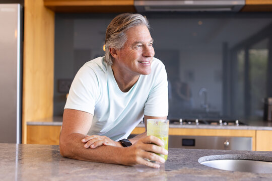 Senior man leaning on kitchen island, holding glass with green smoothie and wearing T-shirt