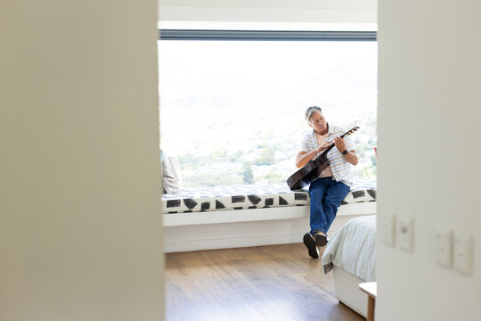 Middle-aged man sitting on window seat playing acoustic guitar in bright bedroom wearing jeans