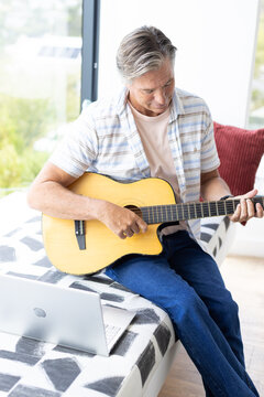 Mature man sitting on patterned bench by window, playing guitar, laptop left, in striped shirt