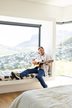 Senior man sitting on built-in window bench playing acoustic guitar beside bed under large window