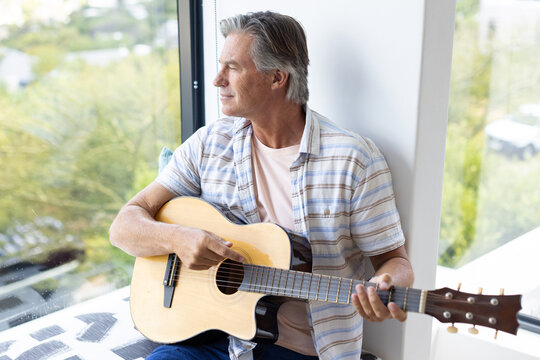 Man sitting on window seat holding acoustic guitar, wearing striped shirt and jeans