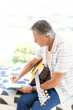 Senior man sitting on patterned bench near bright window, holding guitar and writing in notebook