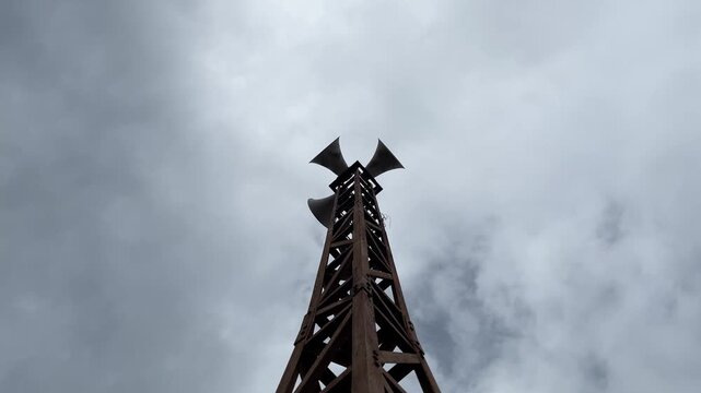 Mosque loudspeaker tower against cloudy daytime sky, viewed from low angle outdoors. Islamic prayer announcement infrastructure creating solemn, spiritual, and atmospheric mood in community setting.