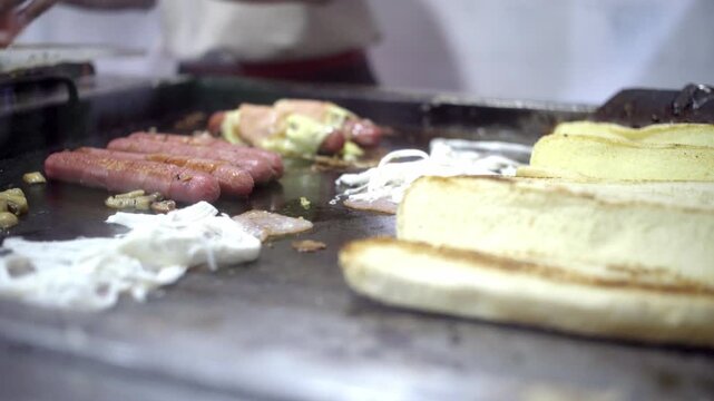 Cooking shot of sausages and bread slices grilling on a hot flat top grill inside a street food stand.