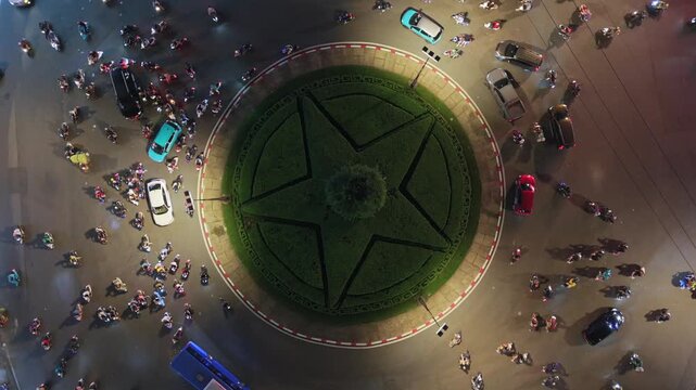 Aerial top-down night view of busy roundabout in Ho Chi Minh City. Motorbikes and cars circle illuminated center, showcasing urban traffic and vibrant nightlife in Vietnam.