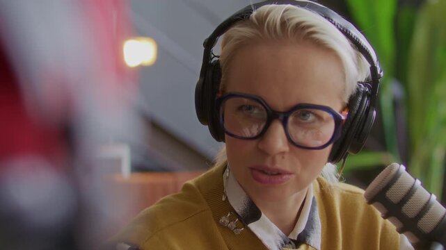 Young woman wearing headphones speaking into microphone during podcast interview recording, smiling and expressing ideas during conversation with host in studio