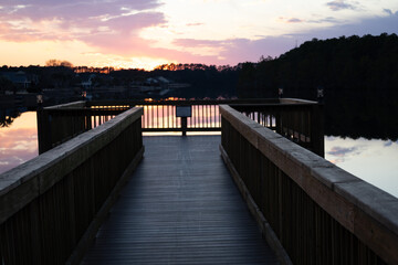 Obraz premium Wooden dock leading into calm lake at sunset with colorful sky reflection and silhouetted trees.